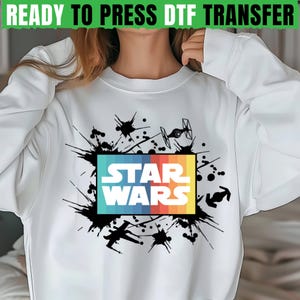 May include: White sweatshirt featuring a colorful "STAR WARS" graphic with a retro design. The design includes a rainbow-colored rectangle and black splatter details. The text "READY TO PRESS DTF TRANSFER" is at the top.