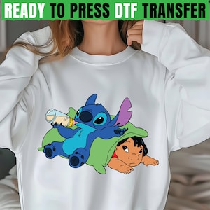 May include: White sweatshirt featuring a cartoon graphic of Stitch drinking from a bottle while lying on a green pillow with Lilo. The text "READY TO PRESS DTF TRANSFER" is at the top.