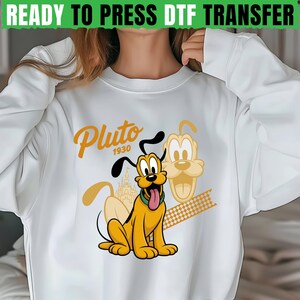 Pode incluir: Camisola branca com um gráfico de desenho animado do Pluto. O design inclui Pluto em amarelo, preto e castanho, com o texto "Pluto 1930" em dourado. As palavras "READY TO PRESS DTF TRANSFER" estão no topo.