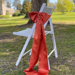Op de afbeelding: Een witte vouwstoel met een grote oranje satijnen strik die om de rugleuning is gebonden.