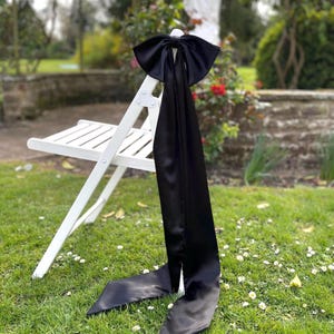 Peut inclure: Une chaise pliante blanche ornée d'un grand nœud en satin noir. Les longues queues du nœud tombent, sur fond d'herbe verte et de feuillage. La chaise est située à l'extérieur, dans un jardin.