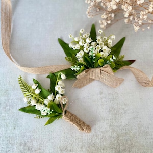 May include: Two small bouquets of white lily of the valley flowers with green leaves, tied with brown twine. One bouquet is tied with a brown bow.