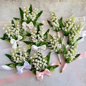 May include: A collection of lily of the valley boutonnieres featuring white bell-shaped flowers and green leaves. Each arrangement is accented with a satin bow in white, cream, or light pink, and some stems are wrapped in white lace or ribbon.