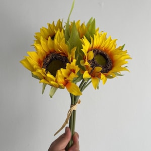 Puede incluir: Un ramo de girasoles artificiales con pétalos amarillos y centros marrones. Los tallos están atados con una cinta marrón.