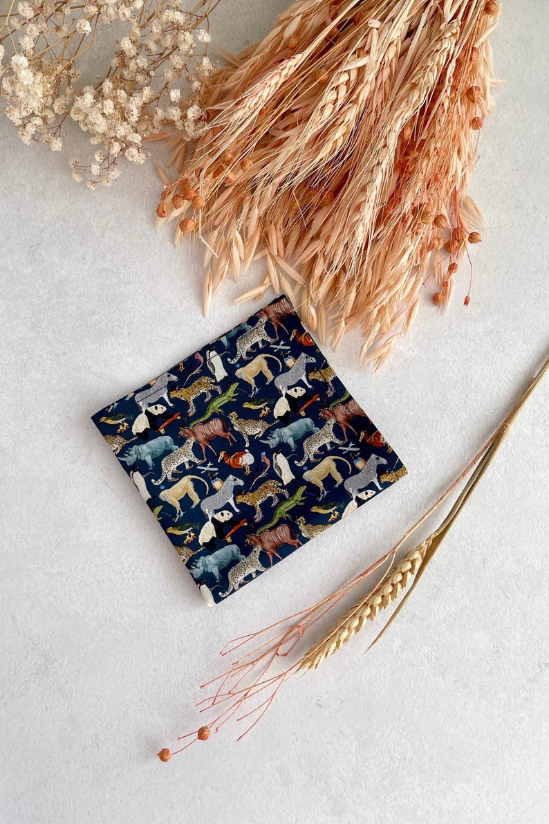 May include: A dark blue fabric square with a repeating pattern of various animals, including zebras, leopards, and rhinoceroses. The fabric is surrounded by dried wheat stalks and white flowers.