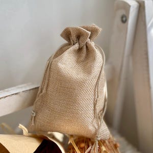 Puede incluir: Una pequeña bolsa de yute de color natural con cordón de ajuste. La bolsa está hecha de un tejido grueso y tejido y tiene un aspecto rústico y hecho a mano.