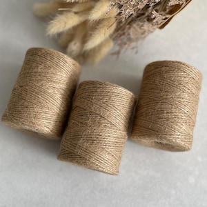 May include: Three spools of natural twine are displayed on a light surface. The twine is tightly wound, and the spools are cylindrical. Dried flowers are visible in the background. Ideal for craft and DIY projects.
