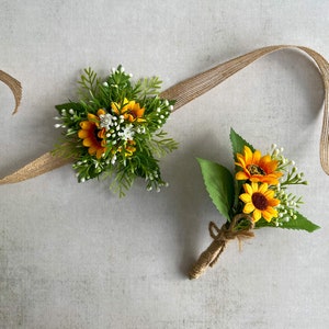 Puede incluir: Dos boutonnières de girasol con cinta de arpillera. La boutonnière más grande tiene tres girasoles y la más pequeña tiene dos girasoles.