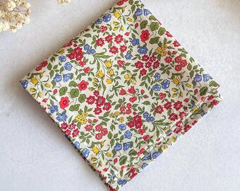 May include: A square fabric pocket square featuring a dense floral pattern on a cream background. The design includes small red, blue, and yellow flowers with green leaves and stems arranged in a repeating, intricate botanical print across the surface.