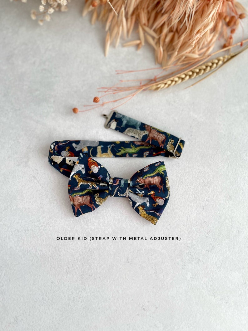 May include: A dark blue bow tie with a colorful animal print pattern. The bow tie has an adjustable strap with a metal buckle. "OLDER KID (STRAP WITH METAL ADJUSTER)" is printed below the bow tie.