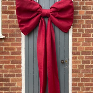 May include: A large, deep red fabric bow with two long, vertical streamers hanging down. The bow is constructed from a textured material and is secured at the centre with a matching fabric knot, creating a full, voluminous shape across the top.