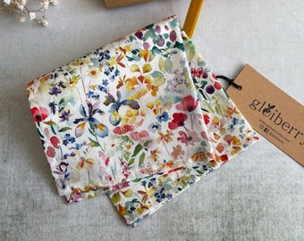 May include: Two white cotton tea towels with a colorful floral pattern. The towels are folded and stacked on top of each other. The pattern features a variety of flowers, including poppies, irises, and daisies. The flowers are painted in shades of pink, red, yellow, blue, and green. The towels are edged with a thin, white trim.