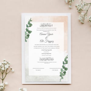 May include: A wedding certificate with the names Amina Yasir and Ali Fayyaz, featuring elegant calligraphy and floral accents. The document is framed by a white border and adorned with green eucalyptus branches and delicate white flowers.