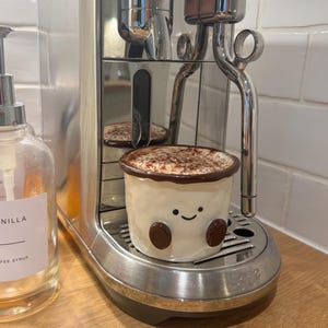 May include: A stainless steel coffee machine with a ceramic mug of cappuccino. The mug is off-white with a smiling face design and brown details. A clear glass bottle of vanilla syrup is to the left.