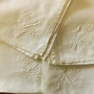 May include: Off-white linen napkins with embroidered floral and geometric designs. The napkins are folded and stacked, showcasing the detailed stitching and delicate patterns. The embroidery features flowers and leaves.