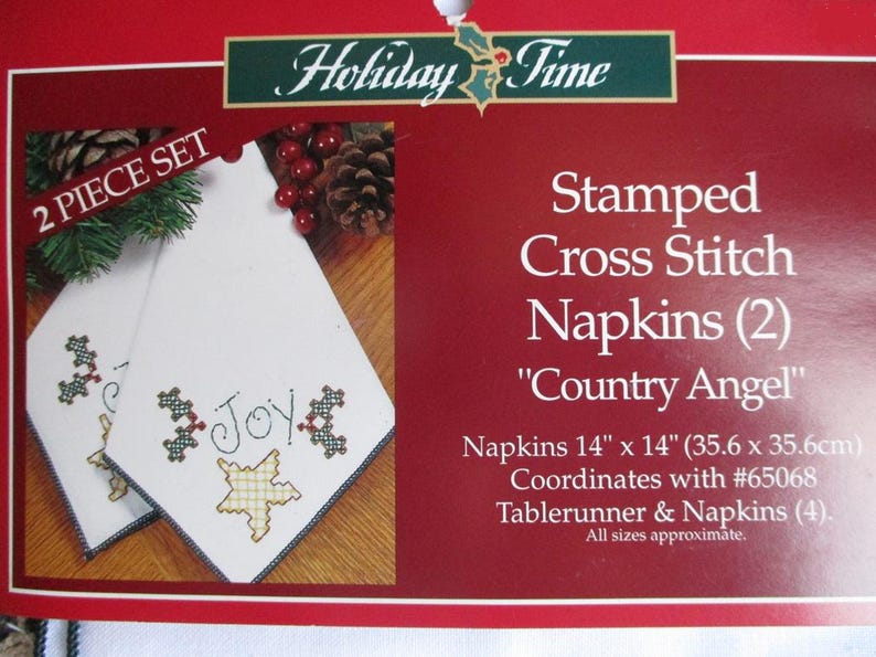 May include: A two-piece set of white cross-stitch napkins with the word "Joy" embroidered on them. The napkins are 14 x 14 inches and feature a "Country Angel" design. The package is red with the words "Holiday Time" at the top.