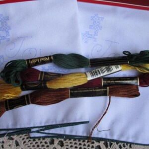 May include: A collection of embroidery floss in various colors, including green, gold, and brown, rests on a white fabric with a blue embroidered design. The floss is neatly arranged, with the spools' labels visible. A green trim borders the fabric.
