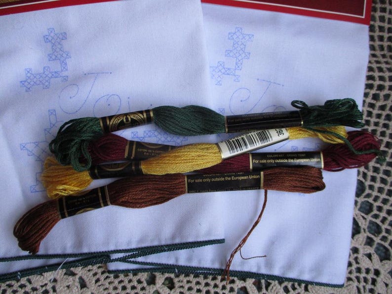 May include: A collection of embroidery floss in various colors, including green, gold, and brown, rests on a white fabric with a blue embroidered design. The floss is neatly arranged, with the spools' labels visible. A green trim borders the fabric.