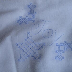 May include: A white fabric with blue cross-stitch designs, including a star, and the word "Joy" in cursive. The cross-stitch patterns are in a pixelated style, creating a unique, handmade aesthetic.