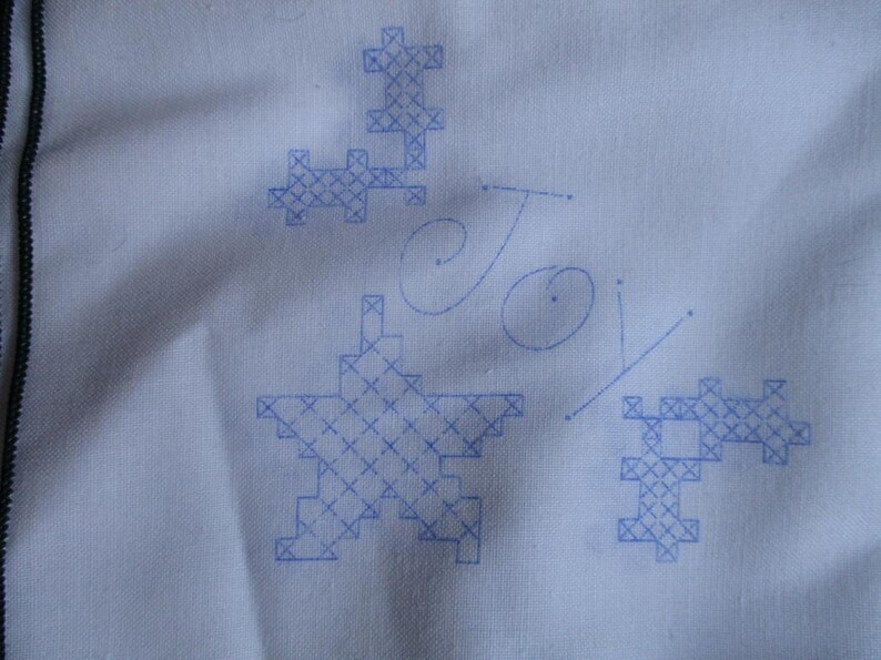 May include: A white fabric with blue cross-stitch designs, including a star, and the word "Joy" in cursive. The cross-stitch patterns are in a pixelated style, creating a unique, handmade aesthetic.