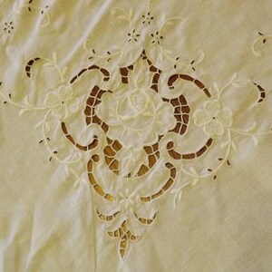 May include: Cream-colored, intricately embroidered tablecloth with a central floral design. The design features a large rose surrounded by cut-out patterns and floral accents. The edges of the tablecloth have a scalloped trim.