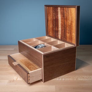 Puede incluir: Una caja de madera para guardar relojes con exterior marrón oscuro e interior de madera clara. La caja cuenta con una tapa abatible, un cajón extraíble y múltiples compartimentos internos para organizar relojes, con un reloj plateado.
