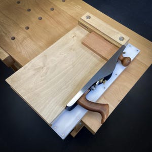 May include: A woodworking plane with a wooden handle and a metal blade is positioned on a wooden workbench. The plane is resting on a light-colored wooden board, with a smaller piece of wood nearby. The workbench has several holes.