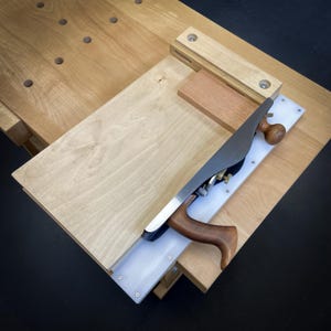 May include: A woodworking plane with a wooden handle and a metal blade is positioned on a wooden workbench. The plane is set up to smooth a light-coloured wooden board. Additional wooden components are also visible.
