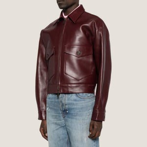 May include: A burgundy leather jacket with a zip-up front, two front pockets, and a collared neckline. The jacket is worn over a pink shirt and paired with blue jeans.
