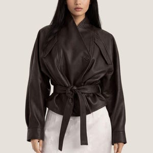 Puede incluir: Chaqueta de cuero marrón oscuro con diseño envolvente. La chaqueta presenta una cintura con cinturón, cierre de un solo botón y mangas largas. El diseño incluye un cuello alto y un detalle único en los hombros.