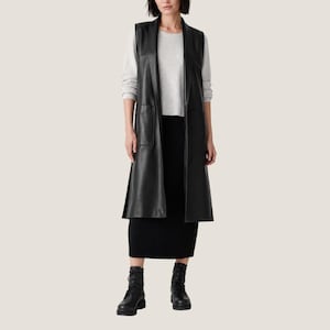 K&ouml;nnte beinhalten: Eine lange schwarze &auml;rmellose Weste mit glatter Oberfl&auml;che und zwei Vordertaschen, getragen &uuml;ber einem hellgrauen Langarmshirt und einem schwarzen Midirock, kombiniert mit schwarzen Schn&uuml;rstiefeln. Die Weste hat ein breites Revers und eine offene Form.