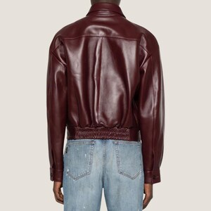 May include: A burgundy leather jacket with a classic collar and buttoned cuffs. The jacket features a ribbed hem and a tailored fit. The model is wearing light blue jeans.