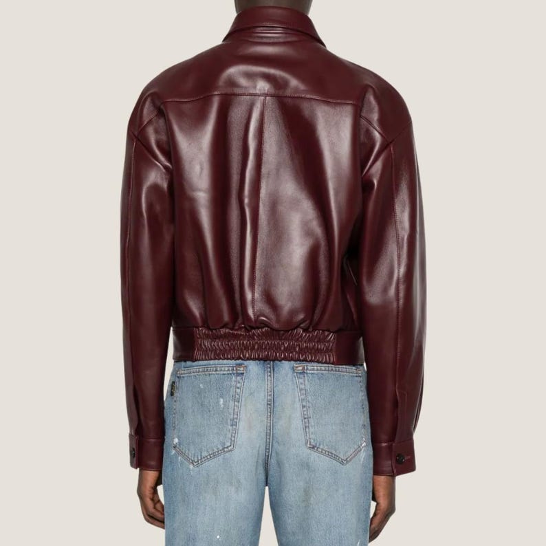 May include: A burgundy leather jacket with a classic collar and buttoned cuffs. The jacket features a ribbed hem and a tailored fit. The model is wearing light blue jeans.