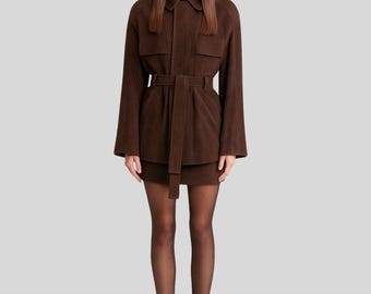 May include: A brown suede jacket with a matching skirt. The jacket has a collared neckline, long sleeves, and a belt at the waist. The skirt is a mini skirt. The person is wearing black sunglasses and black heels.