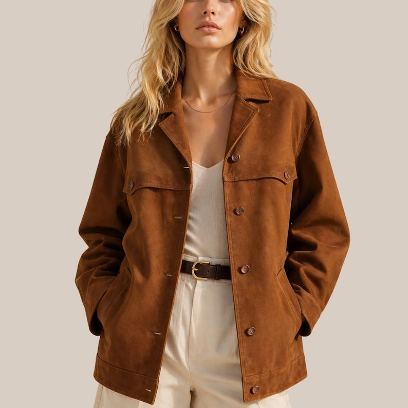 May include: A brown suede jacket with a collared neckline and button-down closure. The jacket features two chest pockets and two side pockets. The model is wearing a white tank top and cream-colored pants. A brown belt with a gold buckle is visible.