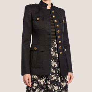 Puede incluir: Una chaqueta negra de estilo militar con botones dorados y cuello alto. La chaqueta tiene dos grandes bolsillos de solapa en la parte delantera y una silueta ajustada.