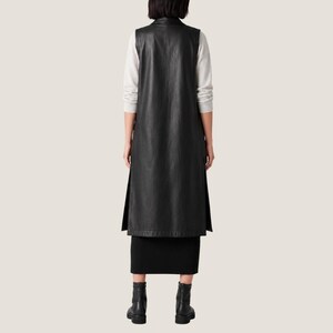 K&ouml;nnte beinhalten: Eine lange schwarze &auml;rmellose Weste aus glattem Material, getragen &uuml;ber einem hellgrauen Langarmshirt und einem schwarzen Rock. Die Weste hat einen hohen Ausschnitt und Seitenschlitze, die vom Saum ausgehen, kombiniert mit schwarzen Stiefeletten.