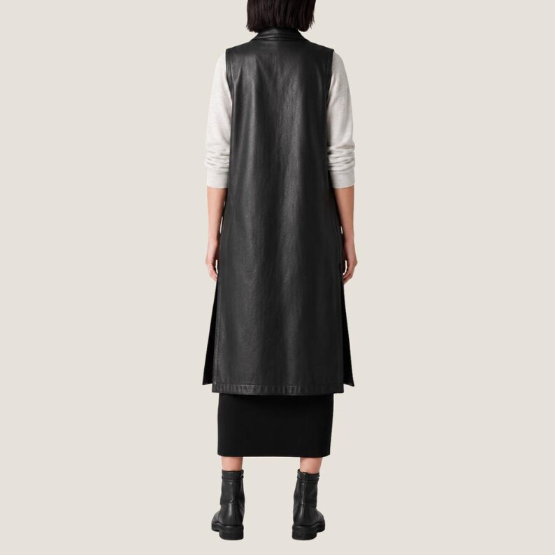 K&ouml;nnte beinhalten: Eine lange schwarze &auml;rmellose Weste aus glattem Material, getragen &uuml;ber einem hellgrauen Langarmshirt und einem schwarzen Rock. Die Weste hat einen hohen Ausschnitt und Seitenschlitze, die vom Saum ausgehen, kombiniert mit schwarzen Stiefeletten.