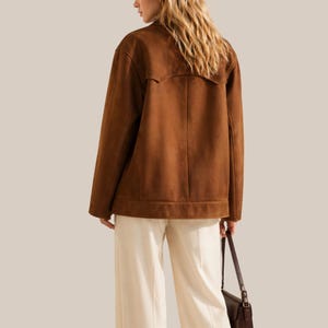 May include: A brown suede jacket with a collar and long sleeves. The jacket is paired with cream-colored pants and a brown leather handbag. The model is facing away from the camera.