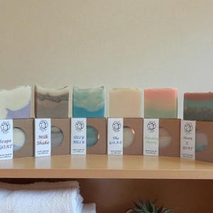 May include: A selection of handmade goat milk soaps in different colours and patterns. Each soap is presented in a brown box with a label and a circular window. The soaps are displayed on a wooden shelf, with a white towel and a small plant in the foreground.
