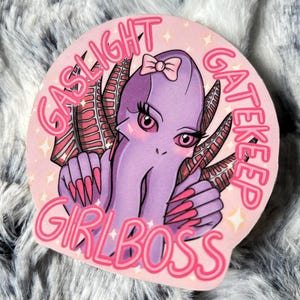 Peut inclure: Autocollant rond avec une illustration de pieuvre de dessin animé. La pieuvre est violette avec des accents roses, de longs ongles roses et un nœud. Le texte "GASLIGHT GATEKEEP GIRLBOSS" est en rose et blanc.