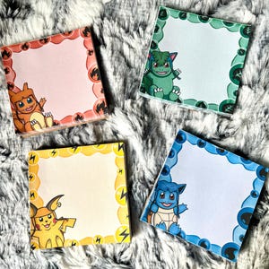 May include: Four square notepads with cartoon character illustrations. Each notepad features a different character with a matching border color: orange, green, yellow, and blue. The notepads are on a fluffy, white and gray surface.