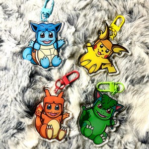 May include: Four acrylic keychains featuring cartoon characters. The keychains depict a blue turtle-like creature, a yellow mouse-like character, an orange lizard-like creature, and a green plant-like creature. Each keychain has a different colored clasp.