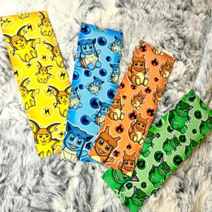 May include: Four colorful rectangular fabric pieces with repeating cartoon character designs. The pieces feature Pikachu on yellow, Squirtle on blue, Charmander on orange, and Bulbasaur on green. Each character is surrounded by related imagery.