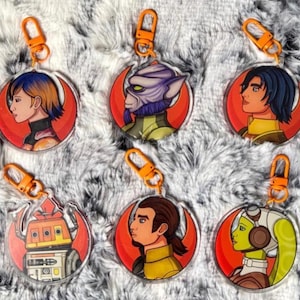 May include: Six circular acrylic keychains featuring illustrated characters from Star Wars Rebels. Each keychain has an orange plastic lobster clasp and displays a character profile against a red crescent background, including a droid and five humanoids.