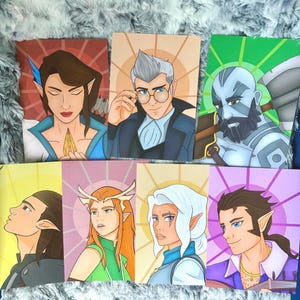 May include: A collection of seven fantasy art prints featuring various characters with detailed facial features and colorful attire. Each print has a unique background color, including red, orange, green, and purple. The characters are depicted with elf-like ears and stylized clothing.