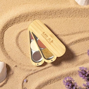 May include: A wooden eyeshadow palette with the word "MILA" printed on the lid. The palette contains six eyeshadow colours, including red, beige, brown, and gold glitter. The palette is open, revealing the colours.