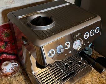 Puede incluir: Una máquina de café espresso Breville de acero inoxidable con acabado de metal cepillado, que presenta una abertura superior para el depósito de granos, un panel de control frontal con botones circulares, un manómetro y una bandeja de goteo.
