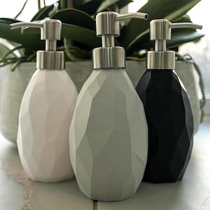 Könnte beinhalten: Drei dekorative Seifenspender in einer Reihe. Die Spender sind in Pastellrosa, Grau und Schwarz gehalten. Jeder hat ein facettiertes, geometrisches Design und eine silberfarbene Pumpe. Im Hintergrund eine Topfpflanze mit grünen Blättern.