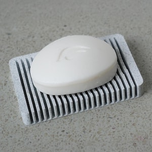 Può includere: Una saponetta bianca con un design rialzato si trova su un portasapone grigio con superficie rigata.